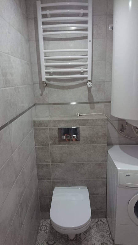 Wykończona łazienka z szarymi płytkami, wiszącą toaletą, grzejnikiem drabinkowym i pralką Amica.