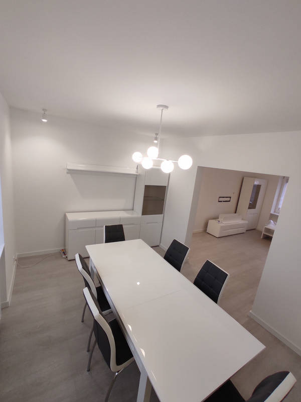 Jasne wnętrze jadalni z białym stołem i krzesłami, widok na salon z białym narożnikiem, nowoczesna lampa sufitowa z kulistymi kloszami, białe ściany i podłoga w odcieniach szarości.