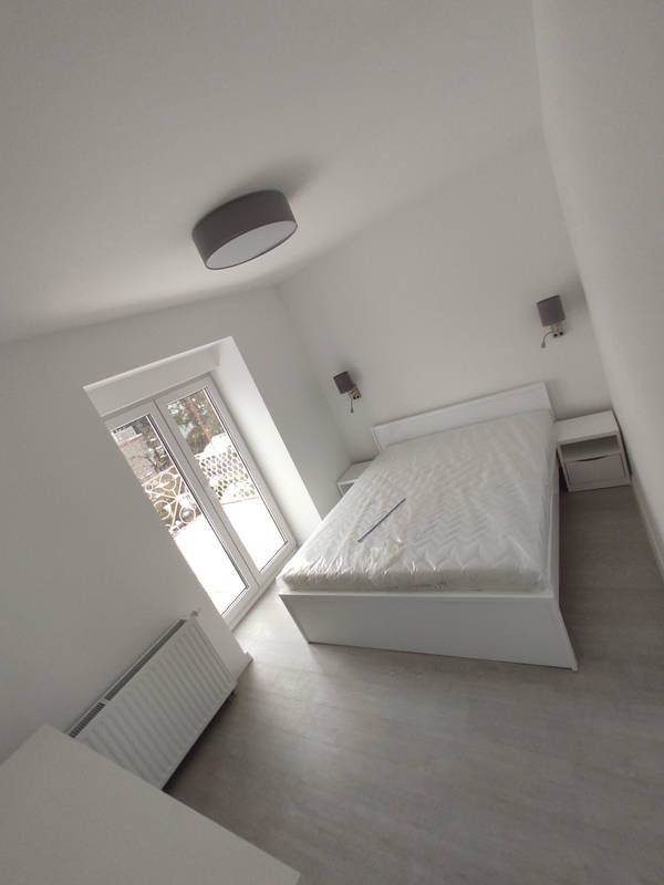 Biała sypialnia z łóżkiem w folii ochronnej, szarymi lampami ściennymi i sufitową, jasną podłogą i grzejnikiem pod oknem.