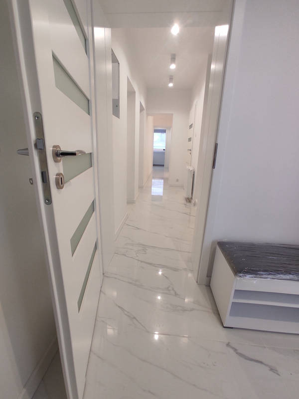 Biały korytarz z marmurową podłogą, widok na wnętrze przez otwarte drzwi, minimalistyczny styl, z widoczną ławką z tworzywa.