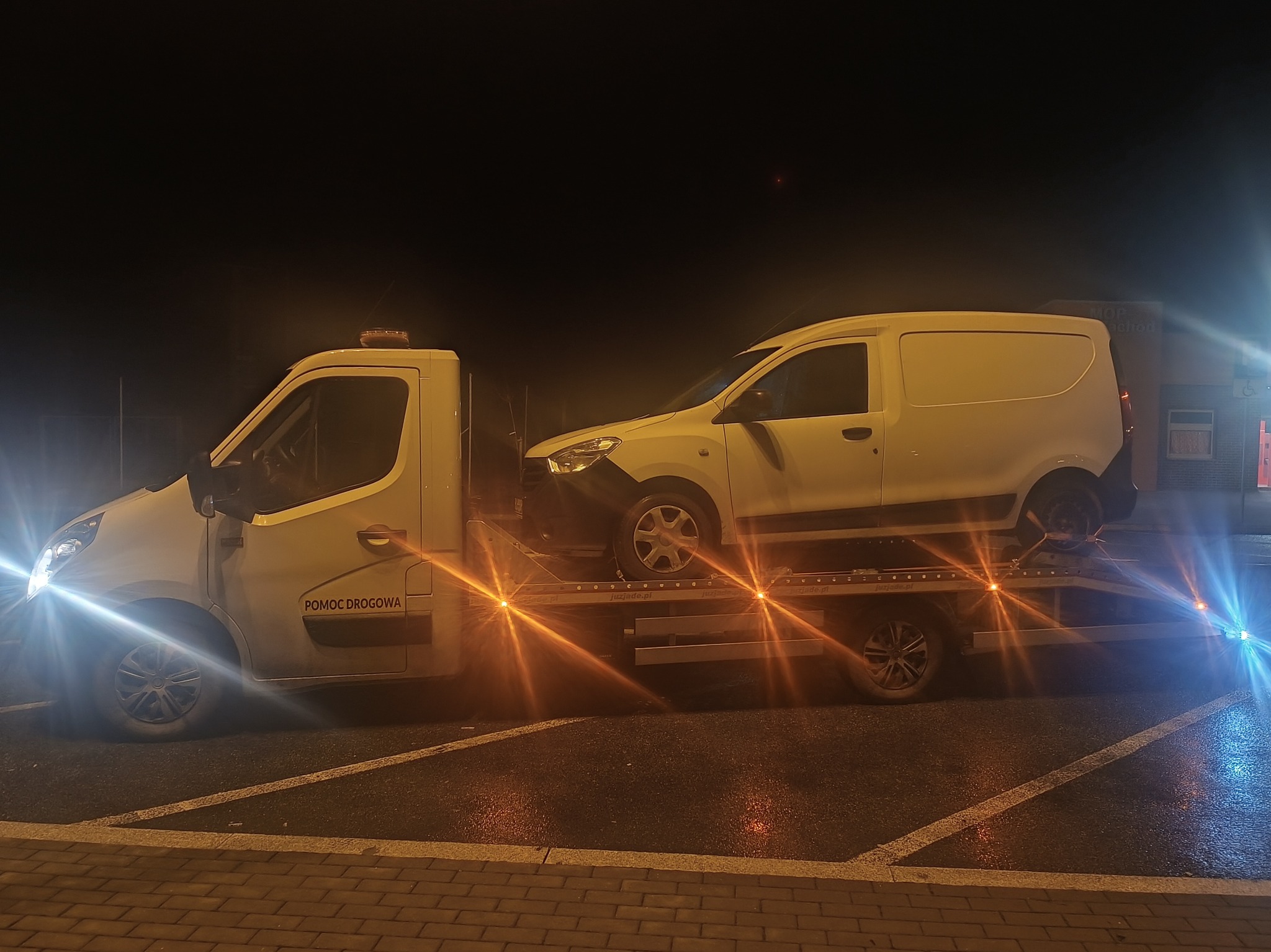 Biała laweta z napisem 'Pomoc Drogowa' przewozi biały samochód dostawczy na tle nocnego parkingu z mokrą nawierzchnią i jasnymi reflektorami.