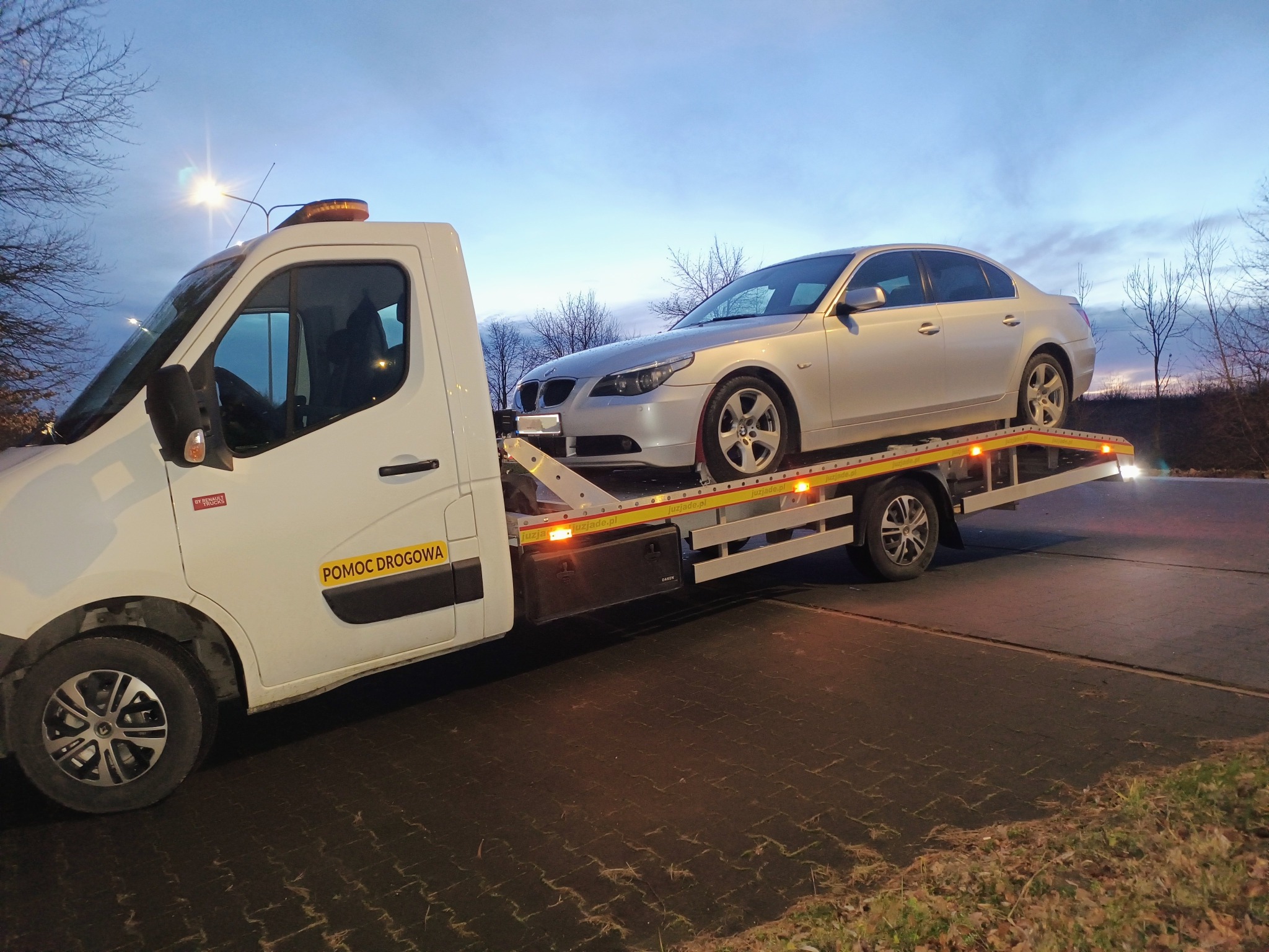 Srebrny samochód osobowy marki BMW załadowany na białą lawetę pomocy drogowej z żółtym napisem 'POMOC DROGOWA' w warunkach zmierzchu.