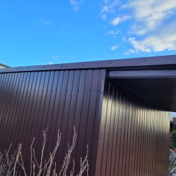 Piotr Wiśniewski - Nowoczesna, brązowa elewacja z blachy trapezowej, widok z dołu na tle błękitnego nieba z chmurami, drewniana konstrukcja w rogu.