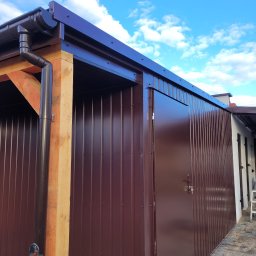 Piotr Wiśniewski - Brązowy blaszany garaż z drewnianą konstrukcją, widoczny system rynnowy i błękitne niebo w tle.