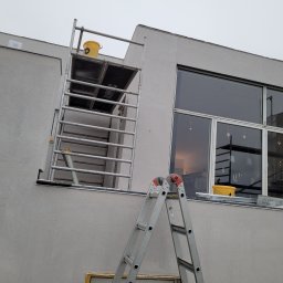 Piotr Wiśniewski - Szarej barwy budynek z dużymi oknami, częściowo zakryty rusztowaniem i drabiną, widoczne ślady świeżo nałożonego tynku.