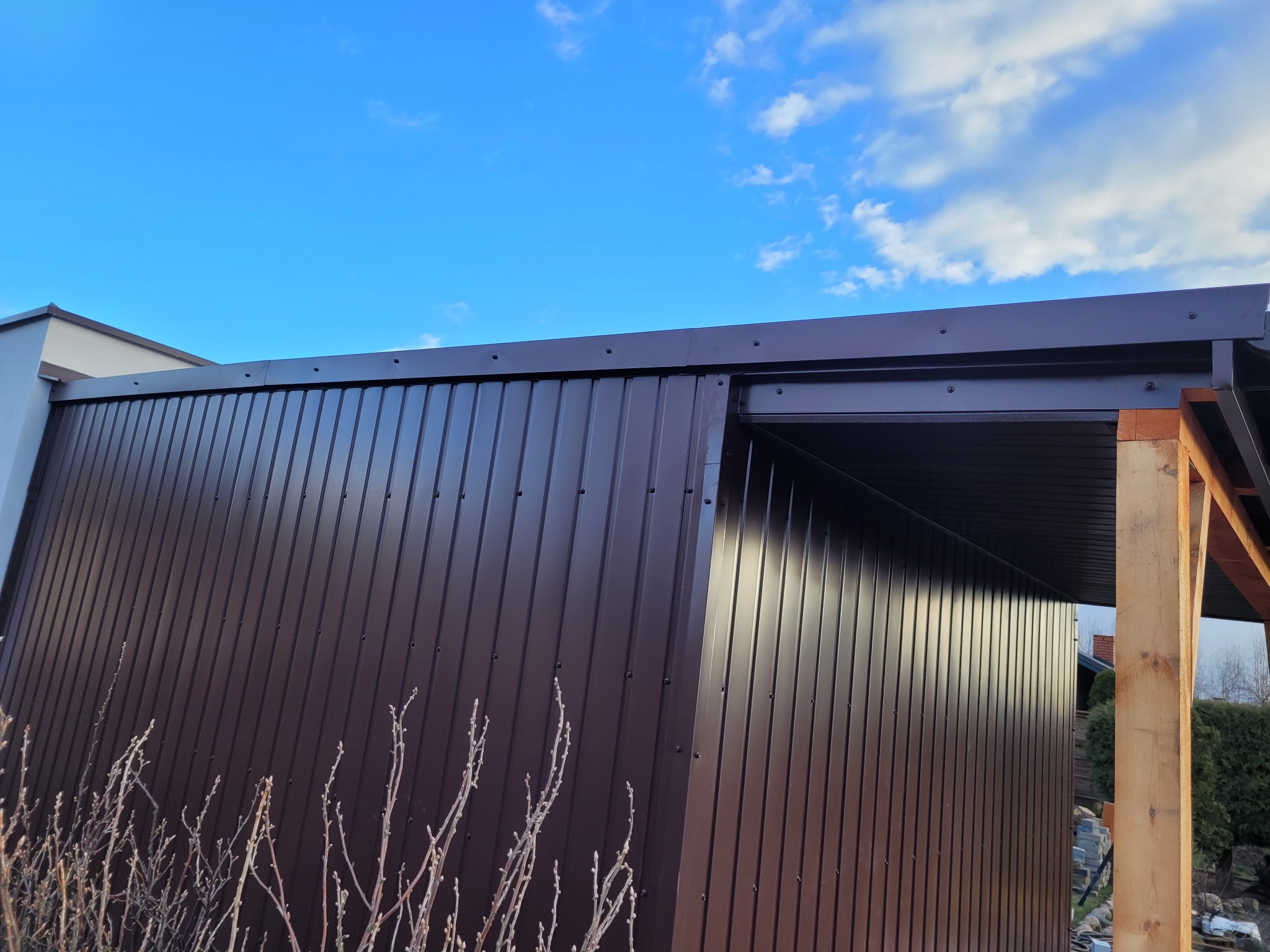 Nowoczesna, brązowa elewacja z blachy trapezowej, widok z dołu na tle błękitnego nieba z chmurami, drewniana konstrukcja w rogu.