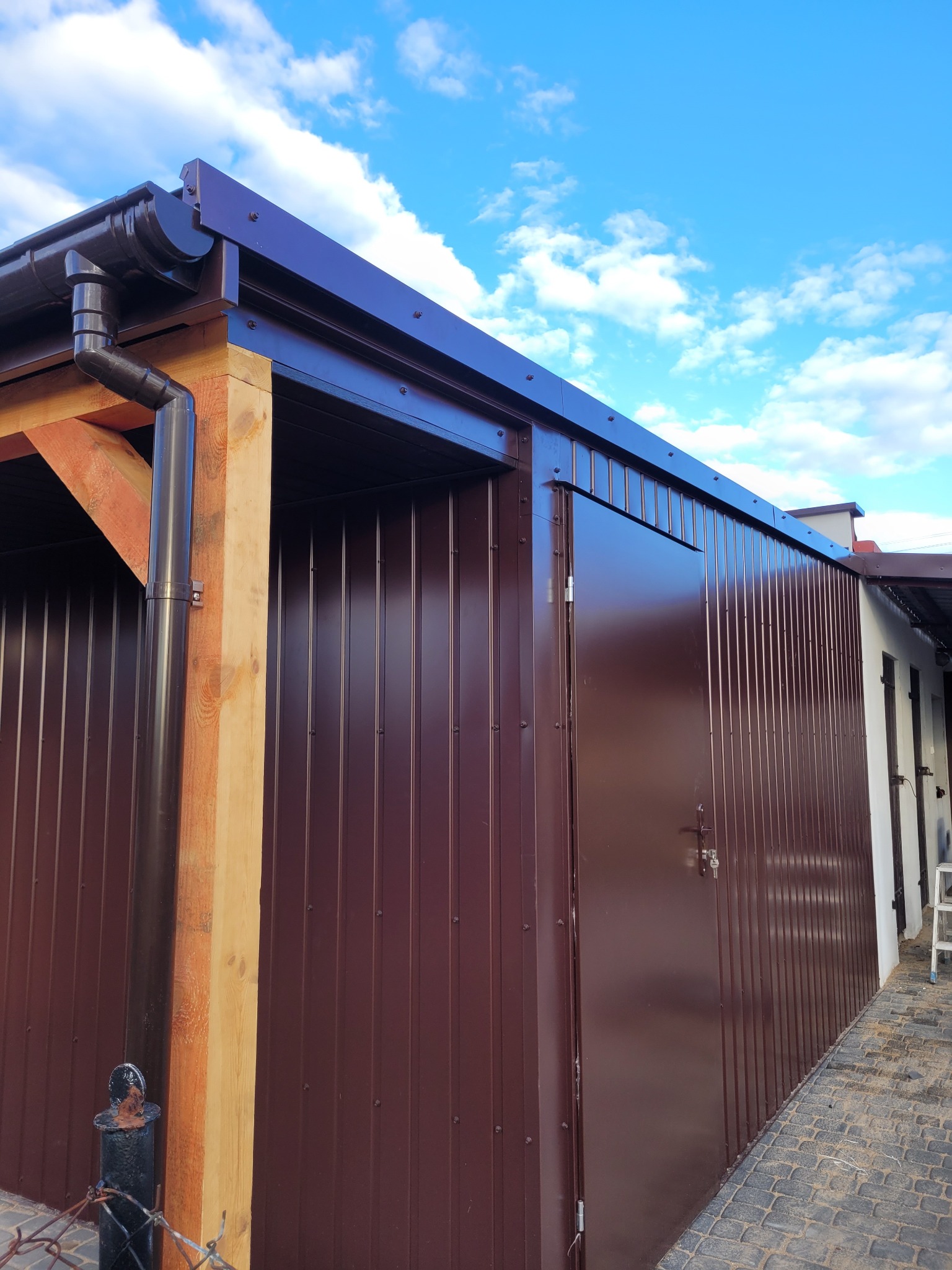 Brązowy blaszany garaż z drewnianą konstrukcją, widoczny system rynnowy i błękitne niebo w tle.