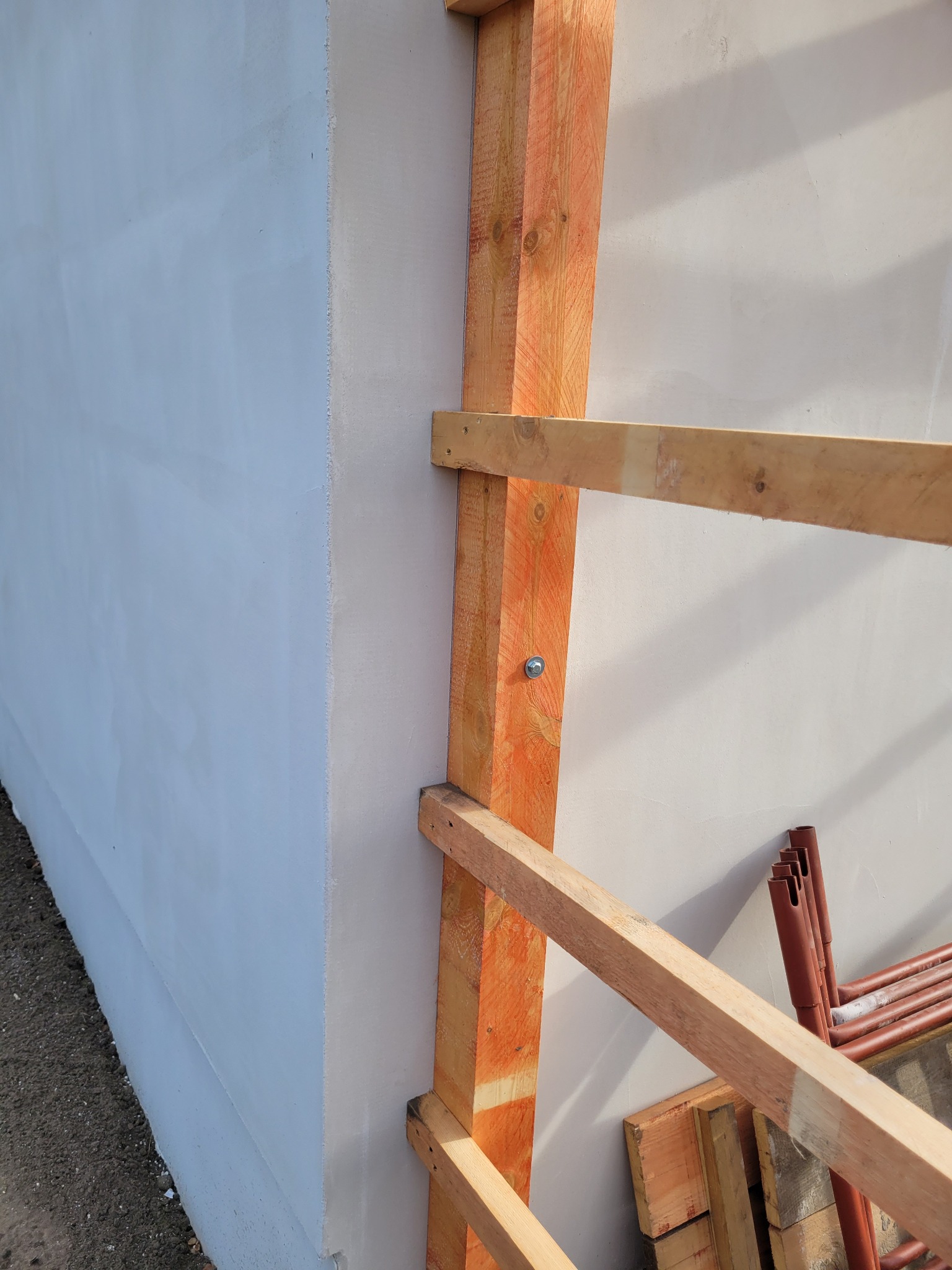 Świeżo otynkowana elewacja budynku z widocznym drewnianym rusztowaniem pomocniczym, przymocowanym śrubami, oraz fragmentem brązowych rur na ziemi.