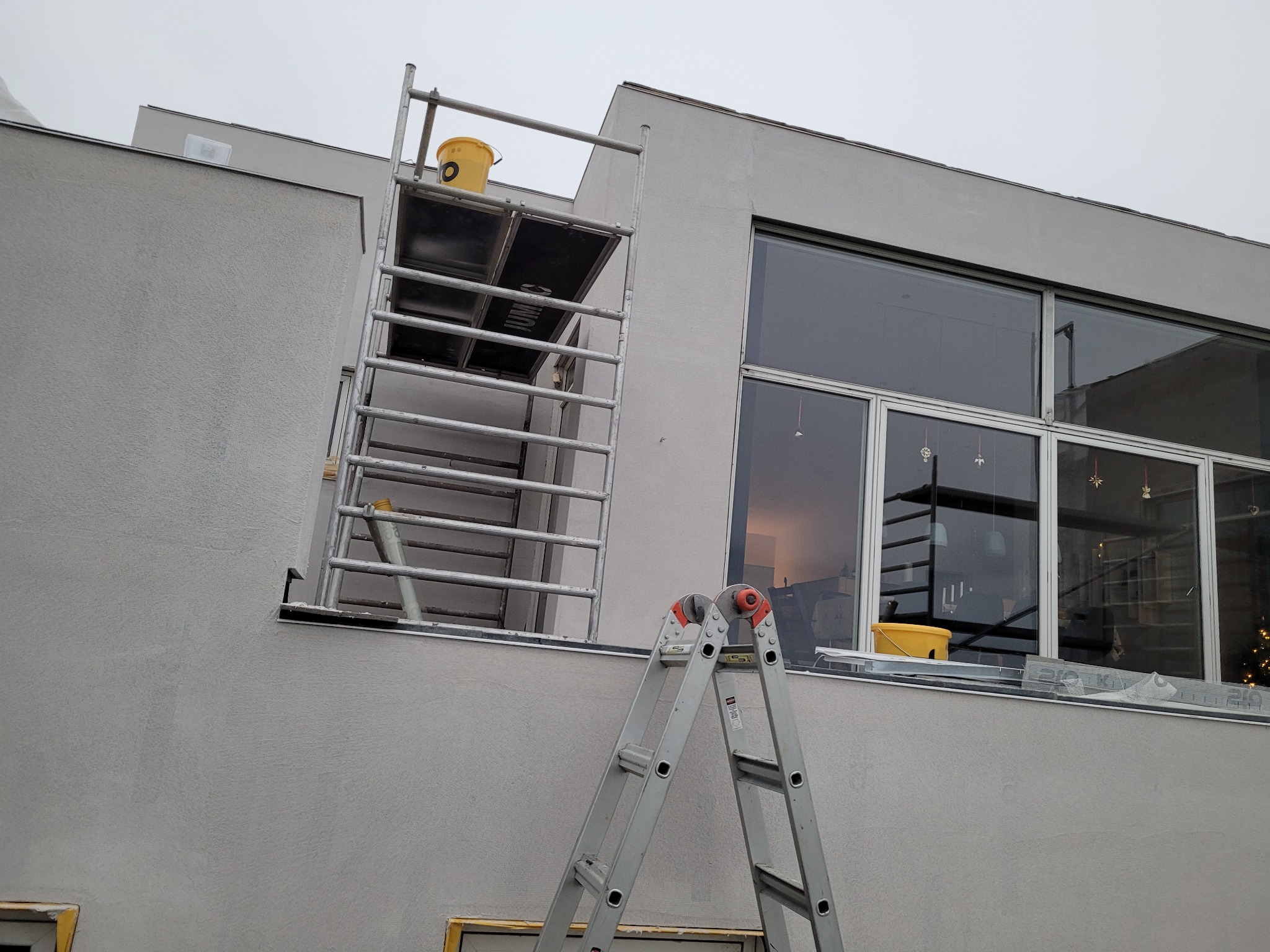 Szarej barwy budynek z dużymi oknami, częściowo zakryty rusztowaniem i drabiną, widoczne ślady świeżo nałożonego tynku.