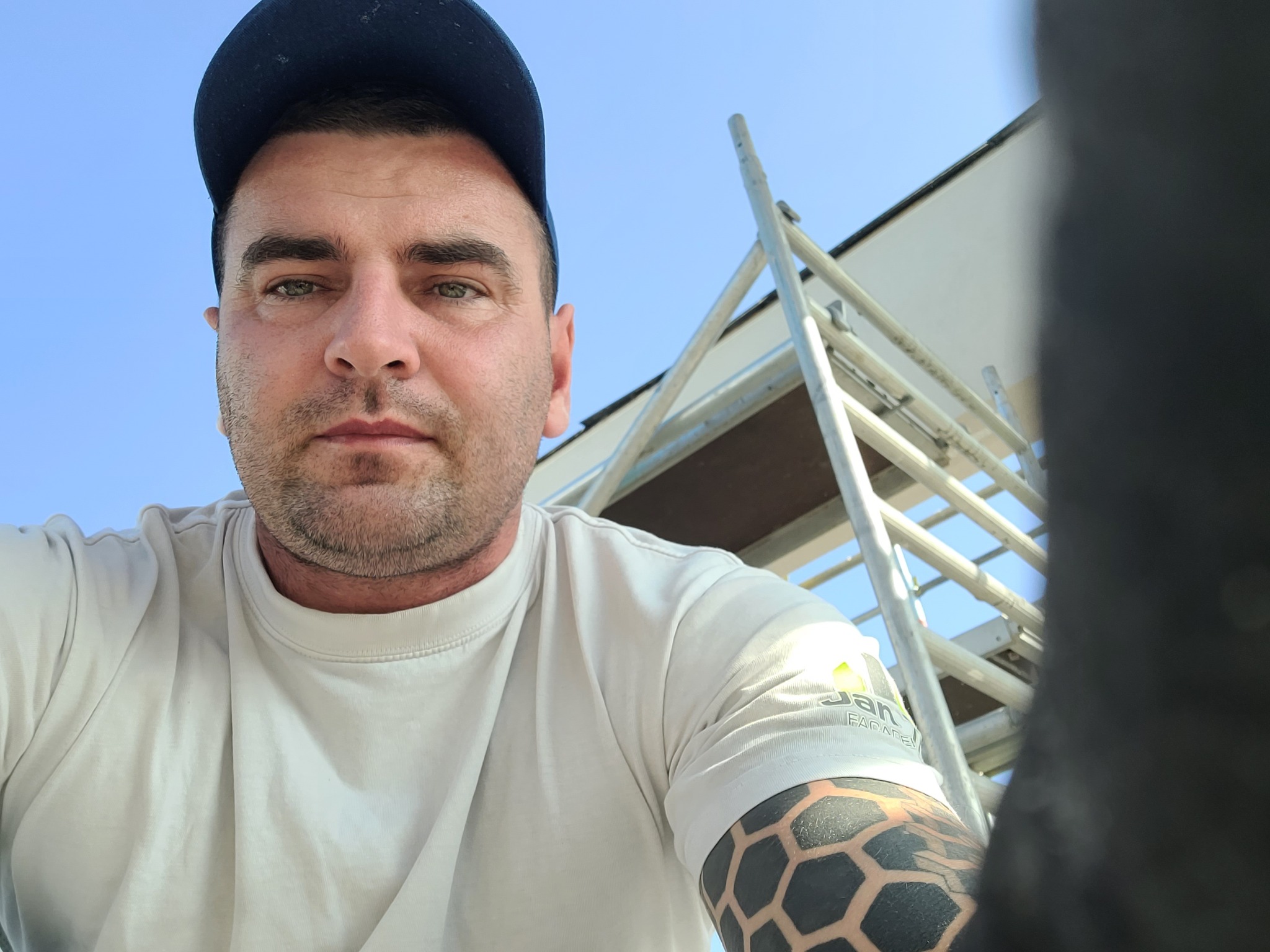 Autoportret pracownika budowlanego w czapce na tle rusztowania i jasnej elewacji budynku, widoczny tatuaż na ramieniu i logo firmy na koszulce.