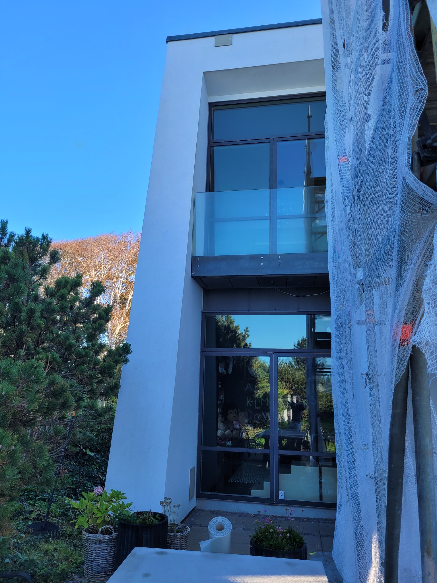 Nowoczesny dom w Kikole z białą elewacją w trakcie prac, widoczne rusztowania z siatką ochronną, okna z balkonem, zieleń w ogrodzie.