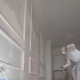 NovaSztuka - Pracownik w kombinezonie ochronnym maluje natryskowo ścianę z dekoracyjnymi listwami w jasnym wnętrzu, widoczne taśmy zabezpieczające przy podłodze.