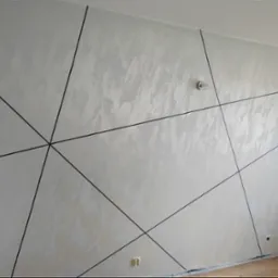 Ściana pokryta dekoracyjną, jasną strukturą z czarnymi, geometrycznymi liniami tworzącymi wzór trójkątów i trapezów, widoczny gniazdko elektryczne i żarówka bez klosza.