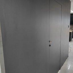 NovaSztuka - Minimalistyczna, ciemnoszara ściana z ukrytymi drzwiami z chromowanym zamkiem, wykończona gładzią i pomalowana, z widoczną jasną podłogą i fragmentem kuchni w tle.