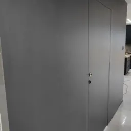 Minimalistyczna, ciemnoszara ściana z ukrytymi drzwiami z chromowanym zamkiem, wykończona gładzią i pomalowana, z widoczną jasną podłogą i fragmentem kuchni w tle.