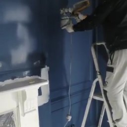 NovaSztuka - Malarz w masce na drabinie szpachluje ścianę w kolorze granatowym w pokoju z ozdobną sztukaterią i zabezpieczonym kominkiem.