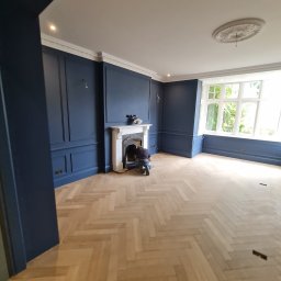 NovaSztuka - Pokój z nową drewnianą podłogą w jodełkę, ciemnoniebieskimi ścianami z dekoracyjnymi panelami i białym kominkiem, przy którym pracuje osoba.