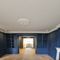 NovaSztuka - Eleganckie, granatowe zabudowy biblioteczne z dekoracyjną sztukaterią i parkietem ułożonym w jodełkę, widok na sąsiednie pomieszczenie z elementami prac remontowych.