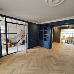 NovaSztuka - Wnętrze w trakcie prac wykończeniowych: parkiet ułożony w jodełkę, zabudowa meblowa w kolorze granatowym, przeszklone drzwi w stylu industrialnym prowadzące na schody, widoczny pracownik w tle.