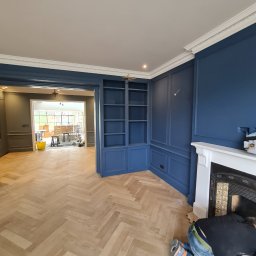 NovaSztuka - Granatowe ściany z dekoracyjnymi panelami i białą sztukaterią, parkiet ułożony w jodełkę, pracownik w jeansach i czarnej koszulce szpachluje okolice kominka.