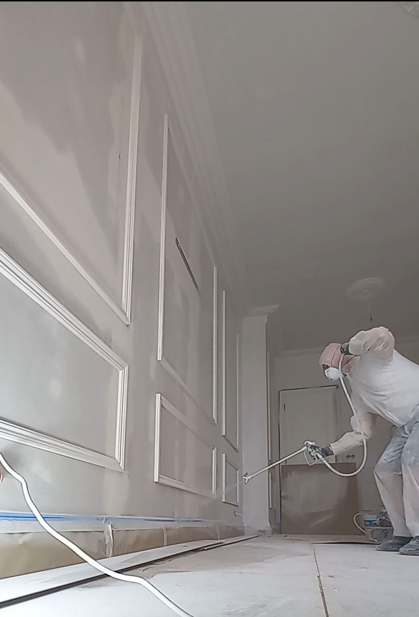 Pracownik w kombinezonie ochronnym maluje natryskowo ścianę z dekoracyjnymi listwami w jasnym wnętrzu, widoczne taśmy zabezpieczające przy podłodze.