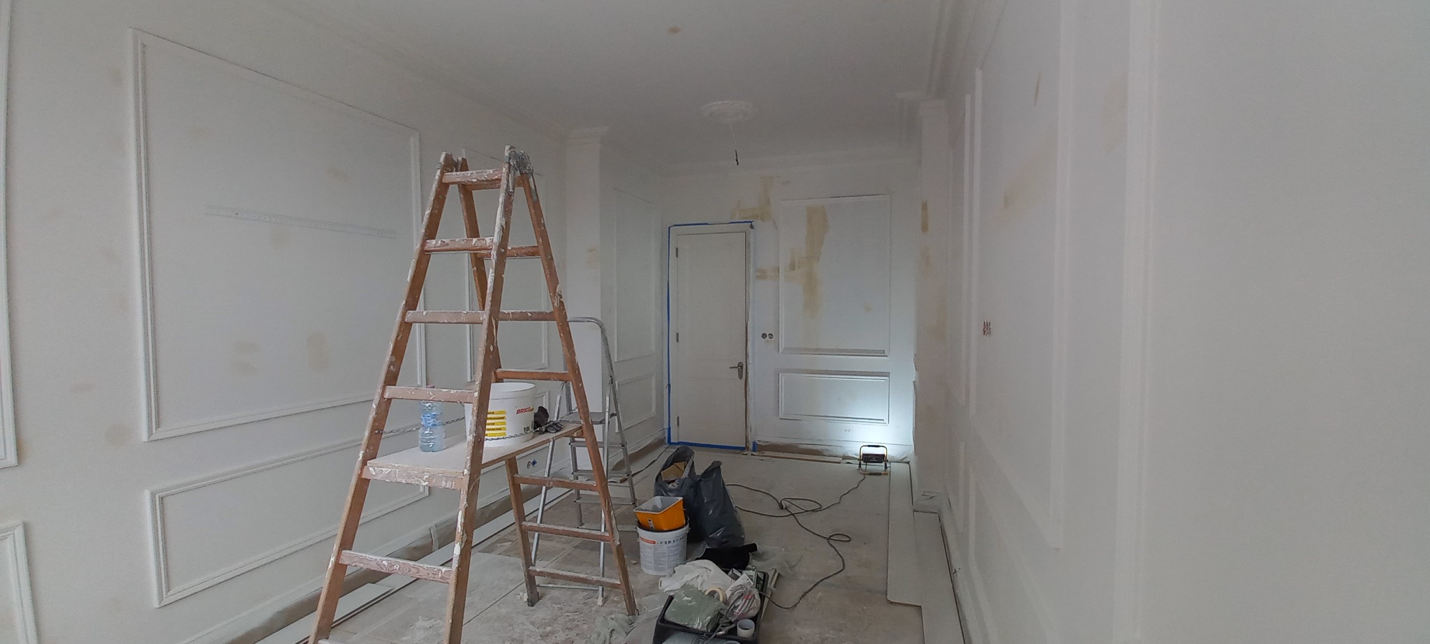 Pokój w trakcie remontu, z drewnianą drabiną, wiadrami z farbą i szpachlą, ściany z dekoracyjnymi listwami w trakcie przygotowania do malowania, zabezpieczone drzwi.
