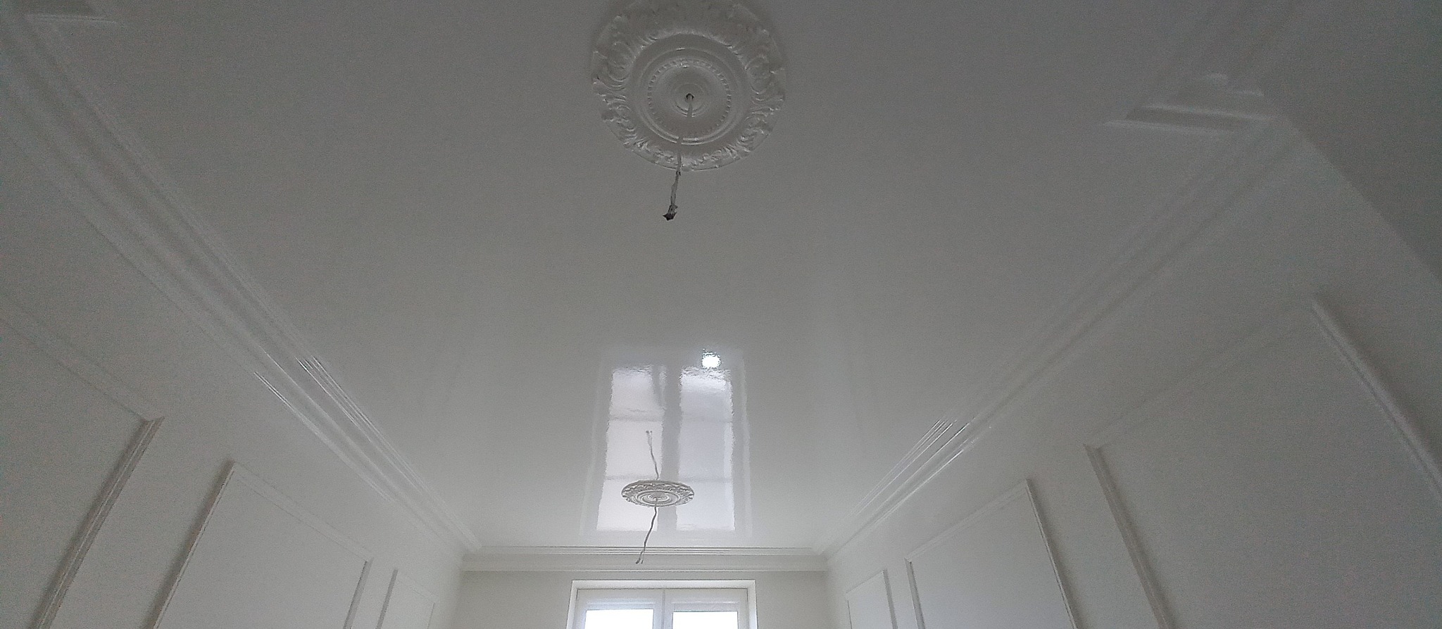 Białe, błyszczące wykończenie sufitu z dekoracyjnym rozetą i listwami, widać odbicie okna, przewody elektryczne wystają z rozety.