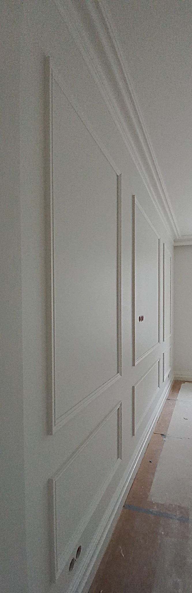 Ściana z dekoracyjnymi panelami i listwami pomalowana na biało, widoczne gniazdka elektryczne w dolnej części ściany, podłoga zabezpieczona kartonem.