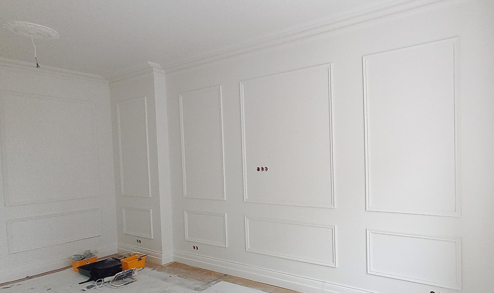 Białe ściany z dekoracyjnymi panelami ściennymi w stylu klasycznym, widoczne puszki elektryczne, częściowo zakryta podłoga z folią malarską i narzędzia malarskie w żółtych wiaderkach.