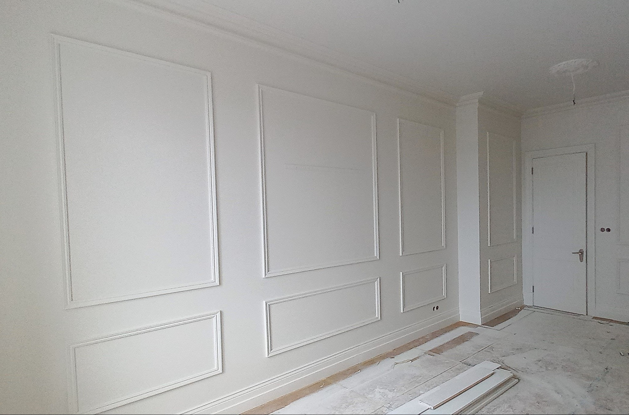 Białe ściany z dekoracyjnymi ramkami i listwami przypodłogowymi w jasnym wnętrzu, widoczne elementy wykończeniowe sufitu i drzwi, podłoga częściowo zabezpieczona folią.
