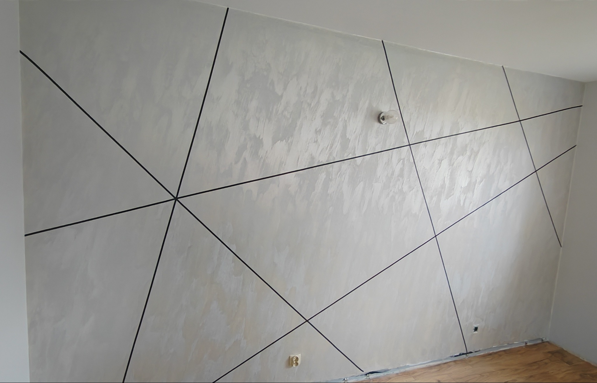 Ściana pokryta dekoracyjną, jasną strukturą z czarnymi, geometrycznymi liniami tworzącymi wzór trójkątów i trapezów, widoczny gniazdko elektryczne i żarówka bez klosza.