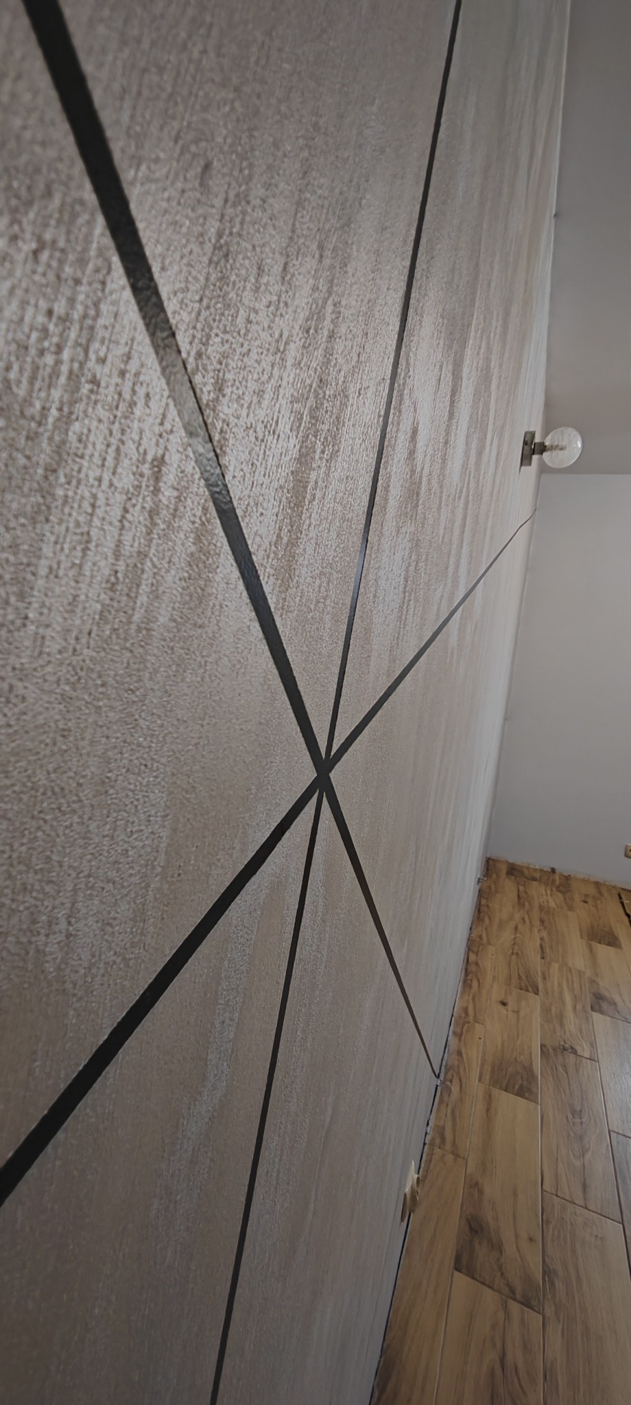 Dekoracyjna ściana z beżową strukturą i czarnymi, geometrycznymi liniami, widziana pod kątem, z fragmentem drewnopodobnej podłogi i kinkietem.