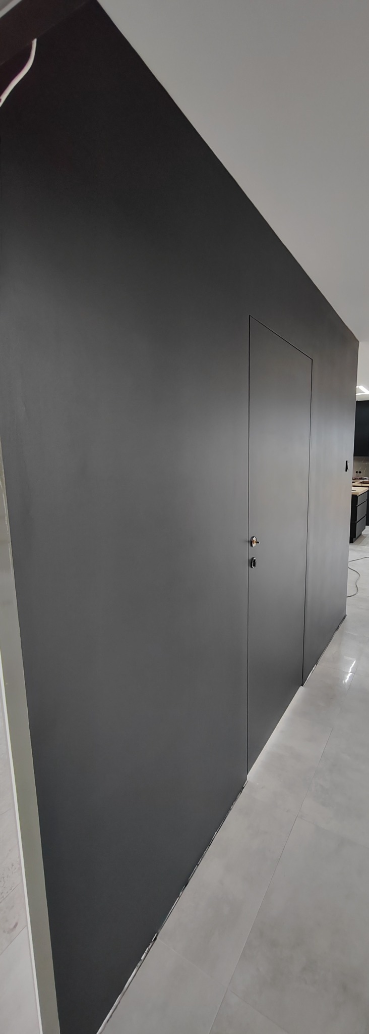 Minimalistyczna, ciemnoszara ściana z ukrytymi drzwiami z chromowanym zamkiem, wykończona gładzią i pomalowana, z widoczną jasną podłogą i fragmentem kuchni w tle.