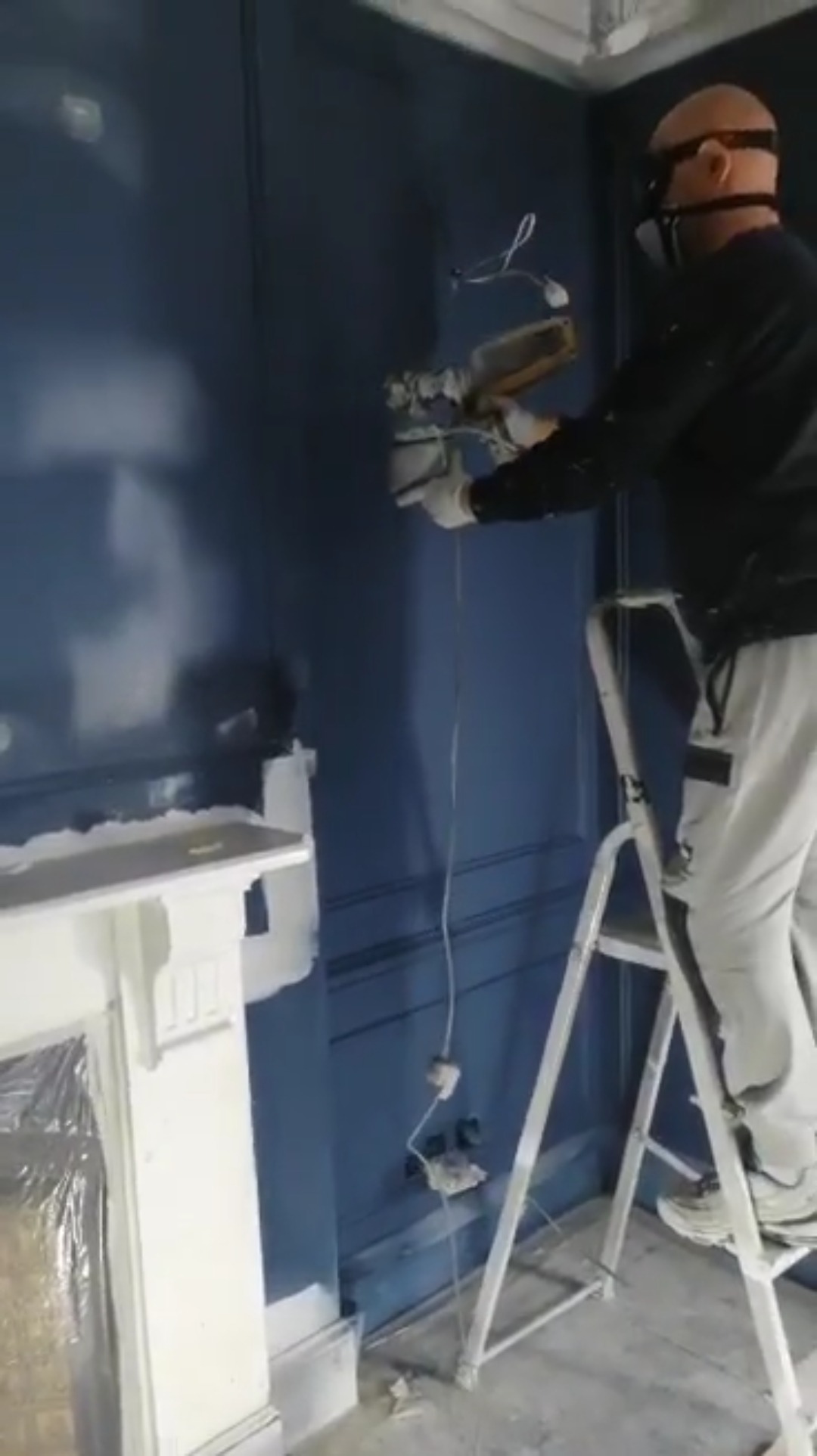 Malarz w masce na drabinie szpachluje ścianę w kolorze granatowym w pokoju z ozdobną sztukaterią i zabezpieczonym kominkiem.