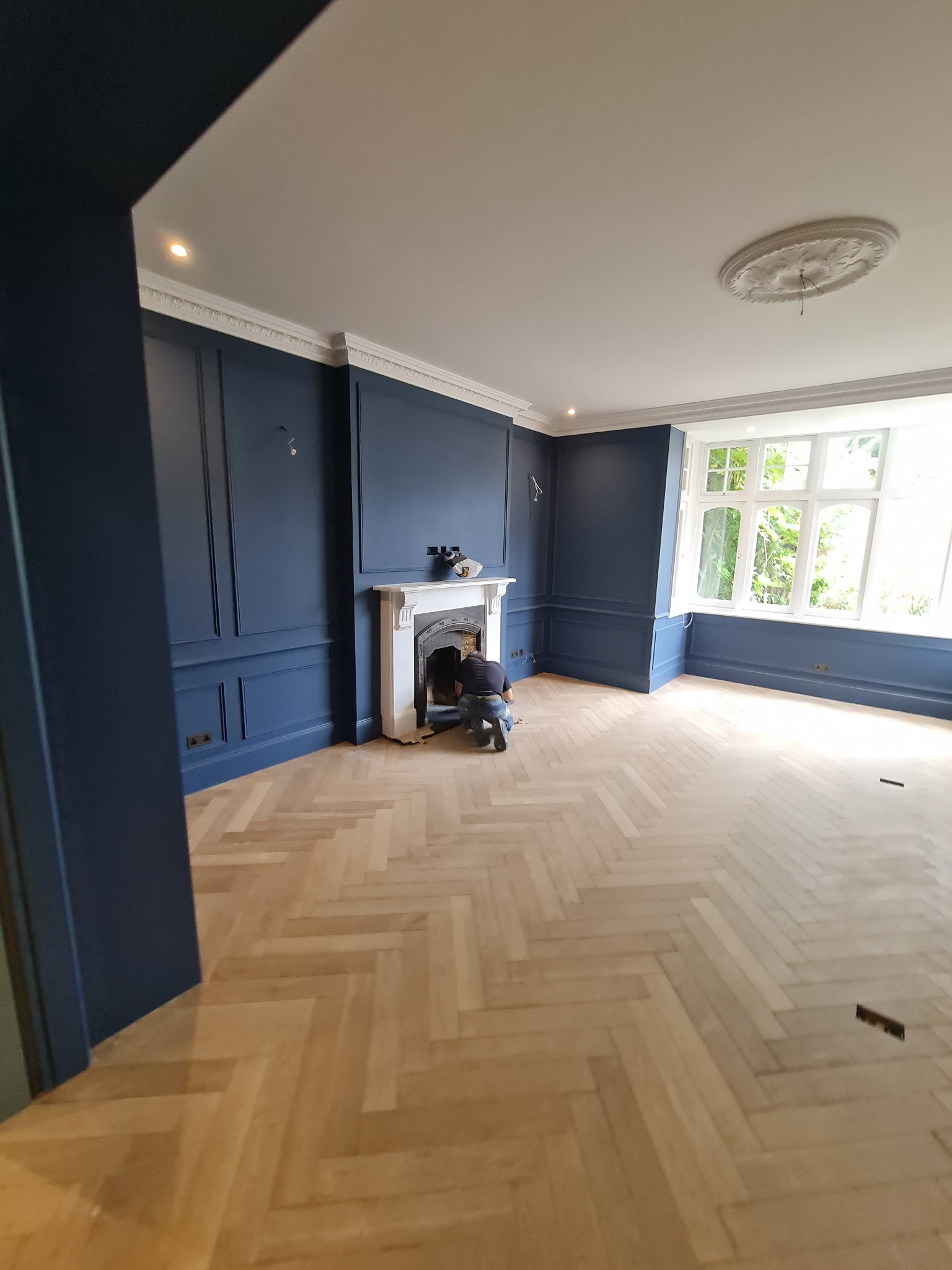 Pokój z nową drewnianą podłogą w jodełkę, ciemnoniebieskimi ścianami z dekoracyjnymi panelami i białym kominkiem, przy którym pracuje osoba.