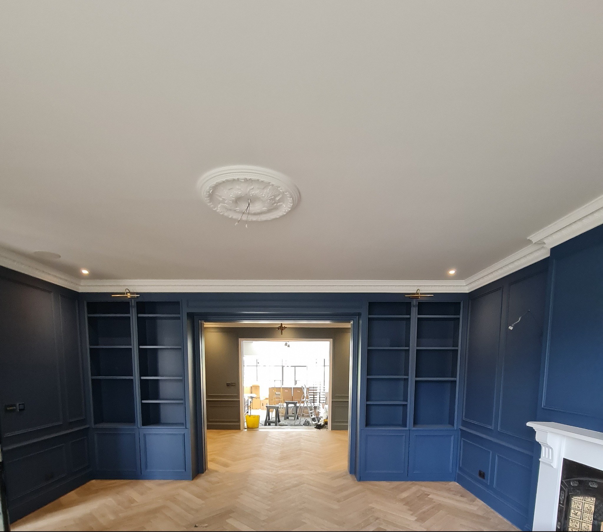 Eleganckie, granatowe zabudowy biblioteczne z dekoracyjną sztukaterią i parkietem ułożonym w jodełkę, widok na sąsiednie pomieszczenie z elementami prac remontowych.