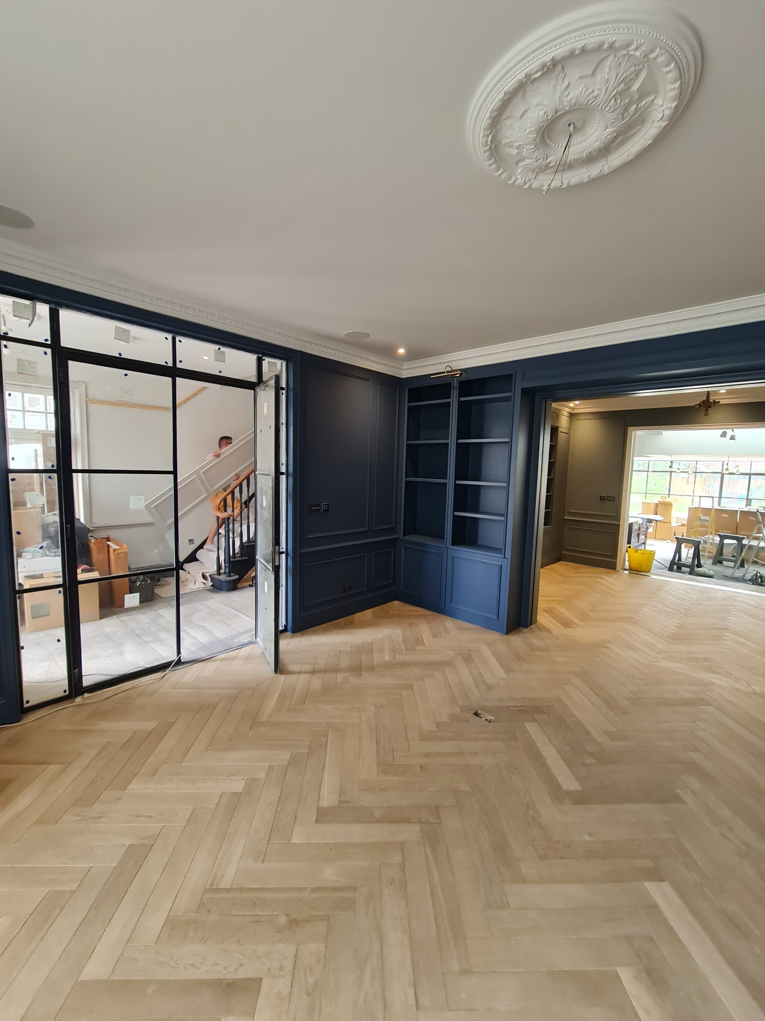 Wnętrze w trakcie prac wykończeniowych: parkiet ułożony w jodełkę, zabudowa meblowa w kolorze granatowym, przeszklone drzwi w stylu industrialnym prowadzące na schody, widoczny pracownik w tle.