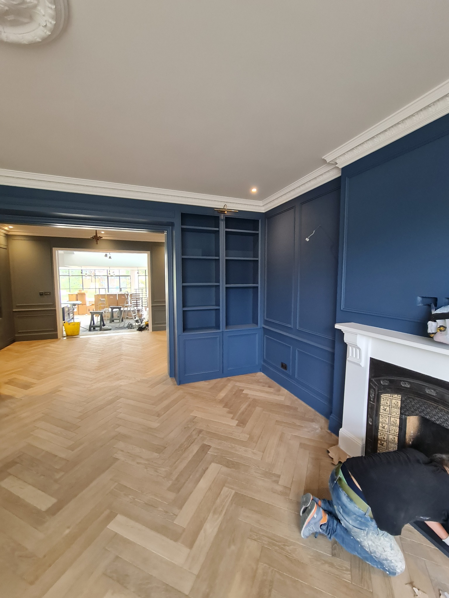 Granatowe ściany z dekoracyjnymi panelami i białą sztukaterią, parkiet ułożony w jodełkę, pracownik w jeansach i czarnej koszulce szpachluje okolice kominka.