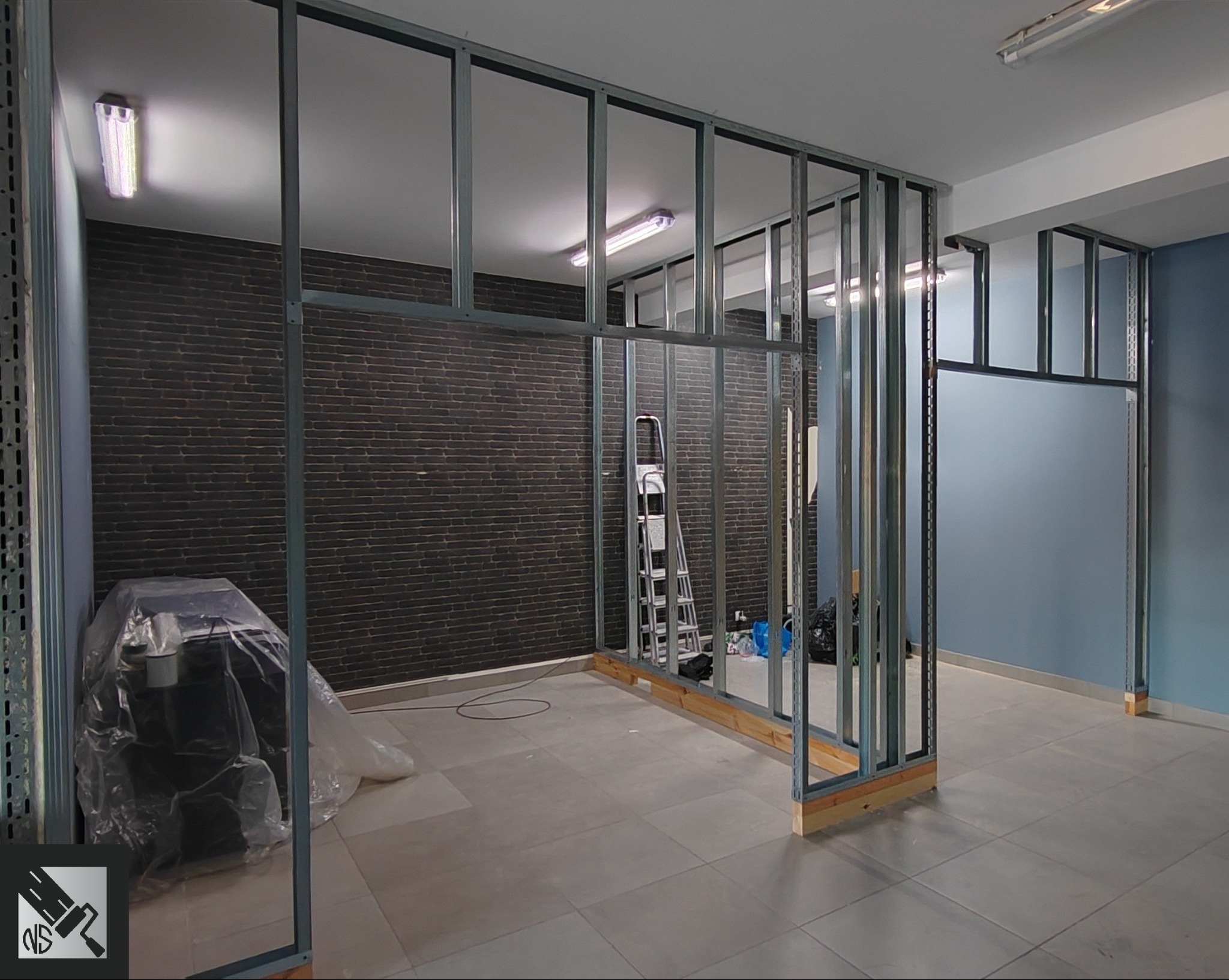 Stelaż z profili stalowych do ścianki działowej w trakcie montażu, widoczna drabina, zakryte folią malarską narzędzia i ceglana ściana w tle.