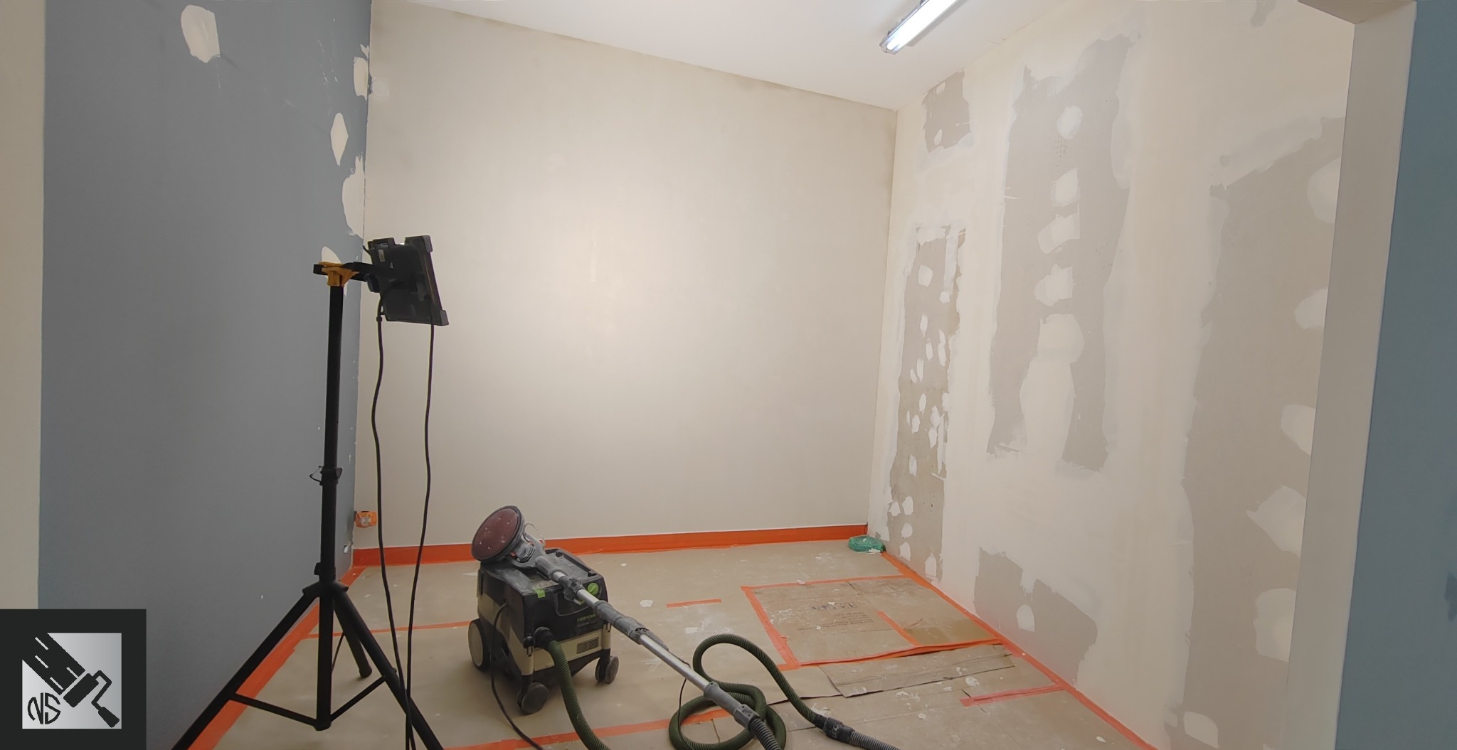 Pokój w trakcie przygotowania ścian do malowania, widoczne ślady szpachlowania i szlifierka z odkurzaczem, podłoga zabezpieczona kartonem.