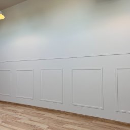 Firma usługowa - Biała ściana z panelami dekoracyjnymi w kształcie prostokątów, drewniana podłoga, oświetlenie sufitowe z okrągłą lampą.