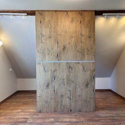 Firma usługowa - Poddasze z drewnianą podłogą i zabudową szafy z przesuwnymi drzwiami o fakturze drewna, oświetlone lampami ściennymi i reflektorami punktowymi.