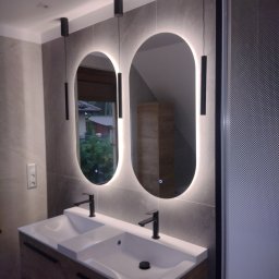 Firma usługowa - Nowoczesna łazienka z podwójną umywalką, owalnymi lustrami z podświetleniem LED i czarnymi, minimalistycznymi bateriami oraz lampami wiszącymi.