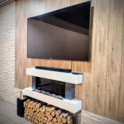 Telewizor na ścianie z drewnianą boazerią, pod nim nowoczesny kominek elektryczny z dekoracyjnym ułożeniem drewna opałowego.