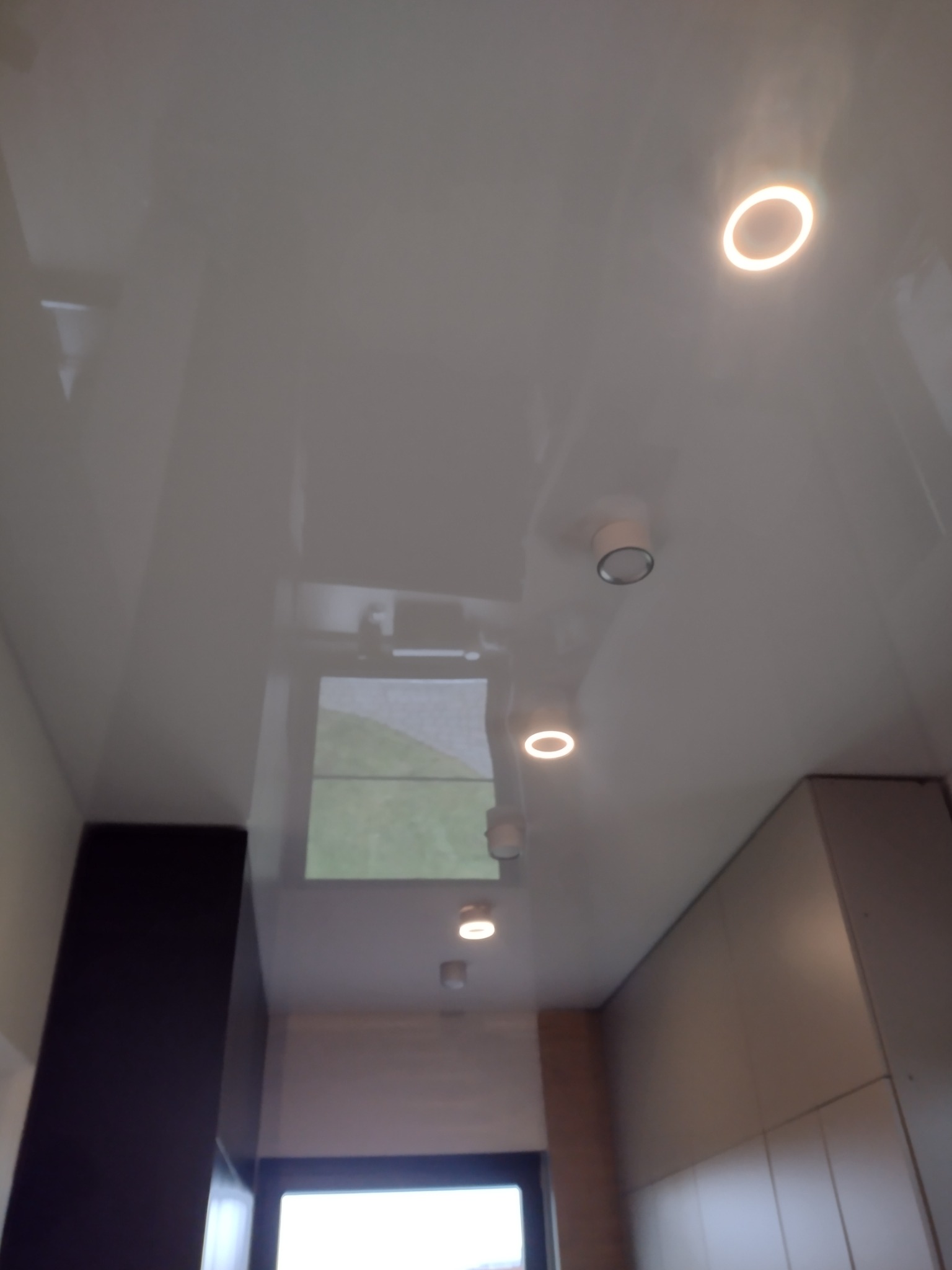 Błyszczący, biały sufit napinany z zamontowanymi okrągłymi lampami LED, widoczne odbicie okna w powierzchni sufitu.