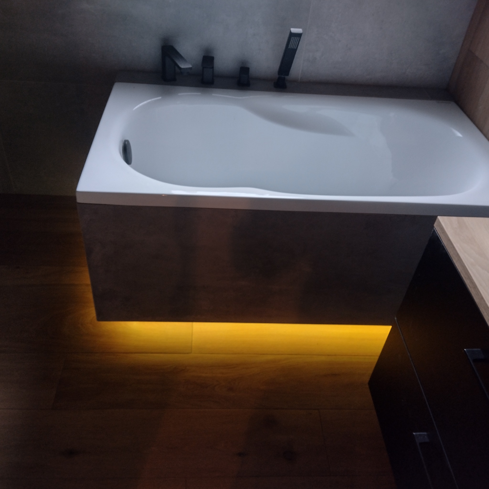 Nowoczesna wanna z czarną armaturą i podświetleniem LED od dołu, na tle szarej ściany i drewnianej podłogi.
