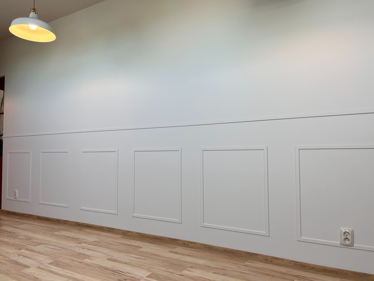 Biała ściana z panelami dekoracyjnymi w kształcie prostokątów, drewniana podłoga, oświetlenie sufitowe z okrągłą lampą.