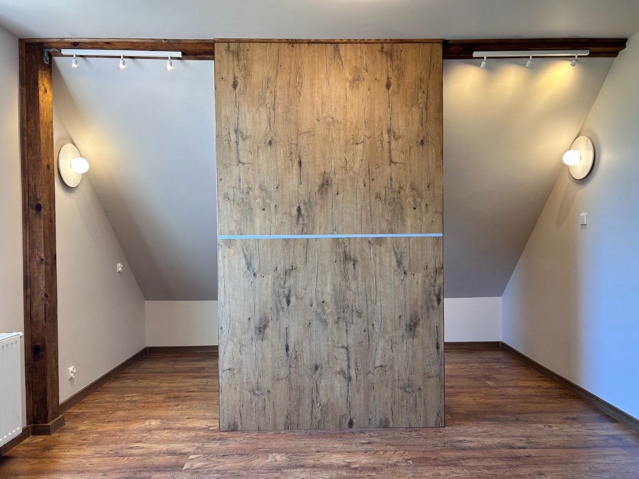 Poddasze z drewnianą podłogą i zabudową szafy z przesuwnymi drzwiami o fakturze drewna, oświetlone lampami ściennymi i reflektorami punktowymi.