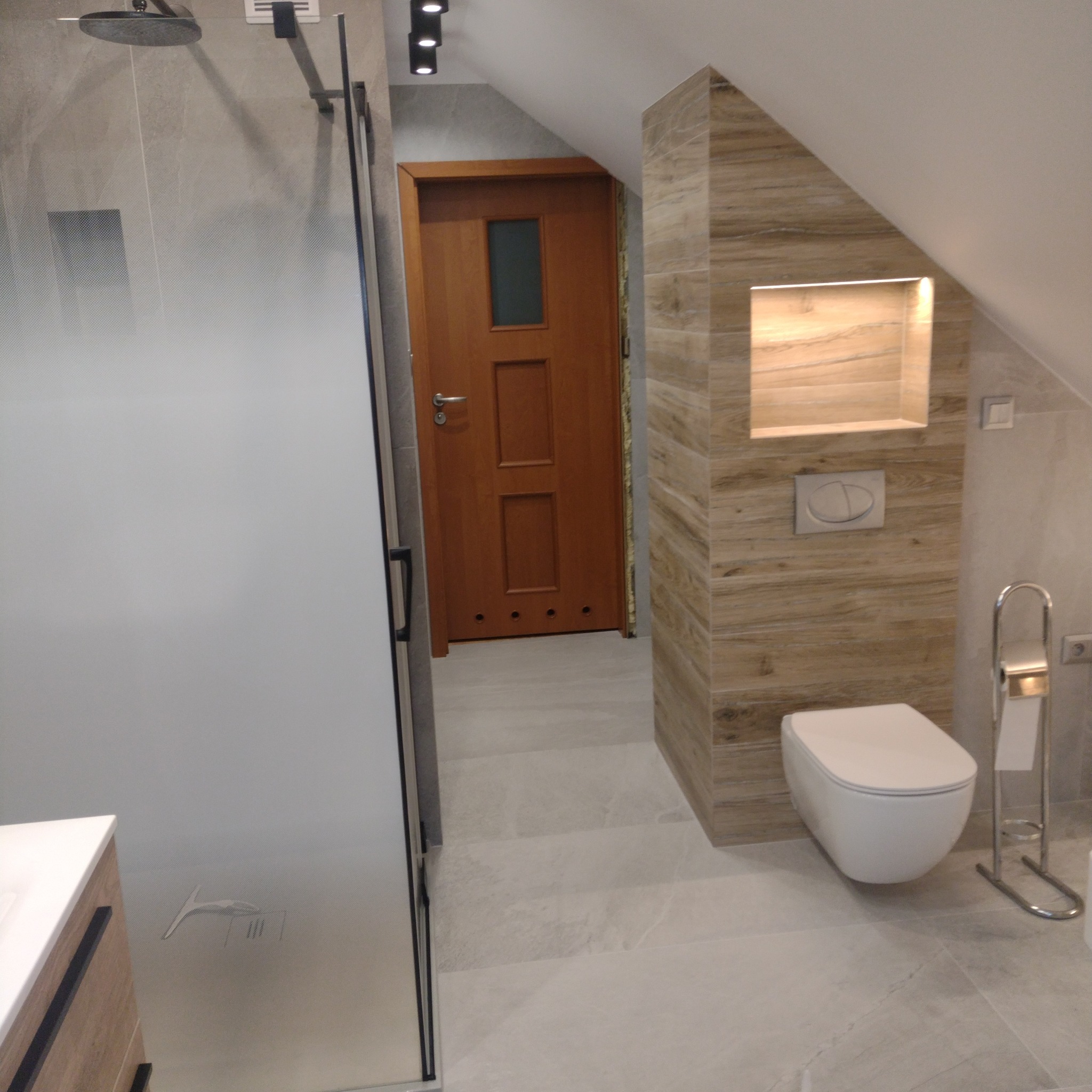 Nowoczesna łazienka na poddaszu z kabiną prysznicową typu walk-in z matową szybą, drewnianą ścianką z podświetloną wnęką i podwieszaną toaletą.