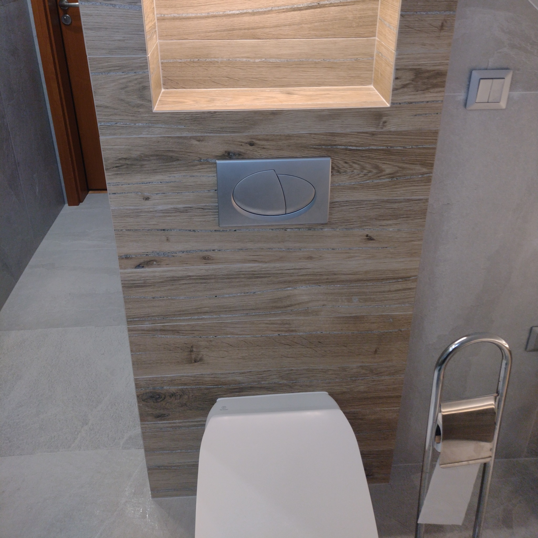Nowoczesna toaleta podwieszana z deską wolnoopadającą, obudowana płytkami imitującymi drewno, z chromowanym przyciskiem spłukującym i uchwytem na papier toaletowy.